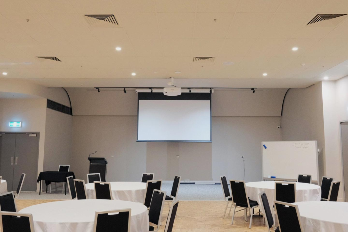 A conference room set up with round tables covered in white cloths and black chairs facing a large projector screen on the stage area.