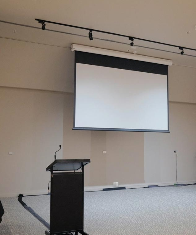 Mobile presentation lectern with microphone in front of a large pull-down projection screen in a conference room edge of a stage area