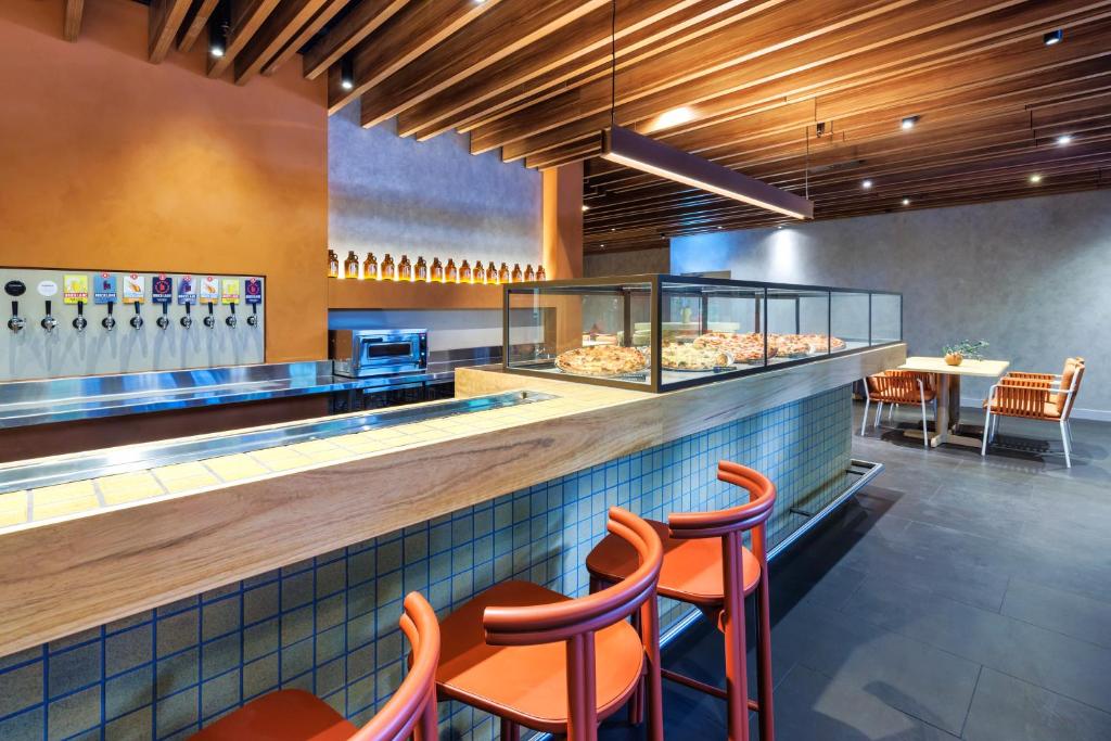 Pizza shop interior with a glass display case of pizzas along a wooden counter and orange bar stools in the foreground.