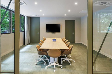 Conference room with a long wooden table and tan swivel chairs, and a wall-mounted TV on the far wall.