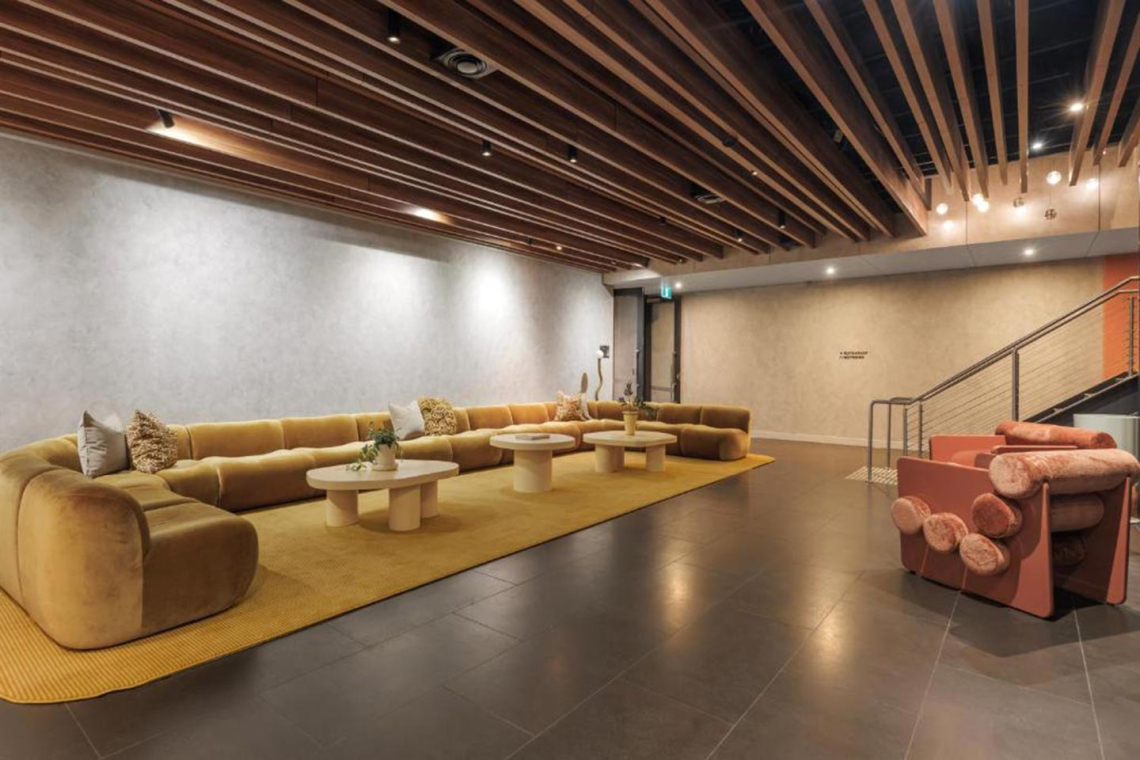 Modern lounge with a long mustard sofa, round coffee tables, warm lighting, and a wood-slatted ceiling; pink seating near the stairs.