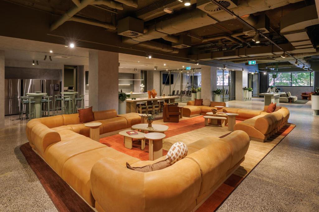 Open-plan lounge with a large orange sectional sofa forming a circle around small coffee tables on a rug; kitchen area visible in the background under an exposed ceiling.