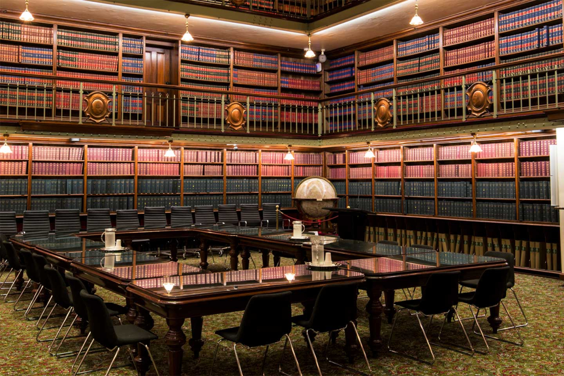 Elegant library conference room with a large wooden table, black chairs, and floor-to-ceiling leather-bound books.