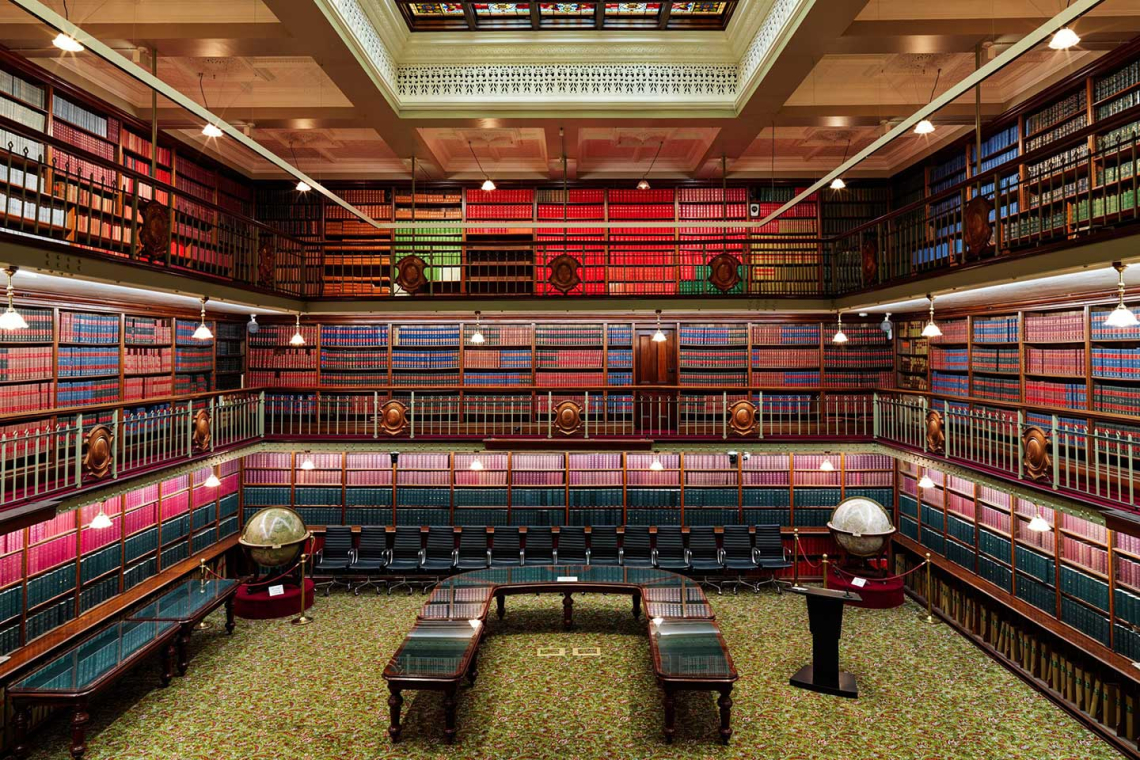 Grand early 20th-century library reading room with two levels of bookshelves, globe displays, and ornate railings surrounding a central carpeted floor.
