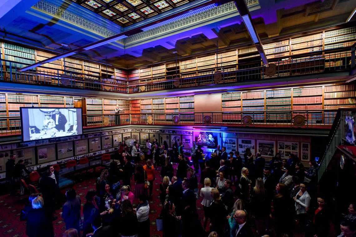 Crowded reception in a grand library with balconies, towering bookshelves, and a large screen showing a black-and-white image.