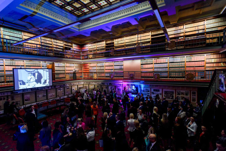 Crowded social event in a grand library-like room with multiple levels of bookshelves and a projector screen at the center.