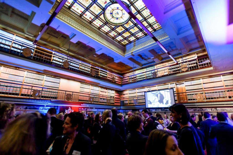 Guests mingle at a formal event in a grand library with a stained glass skylight and tall bookshelves, a projector screen in the background.