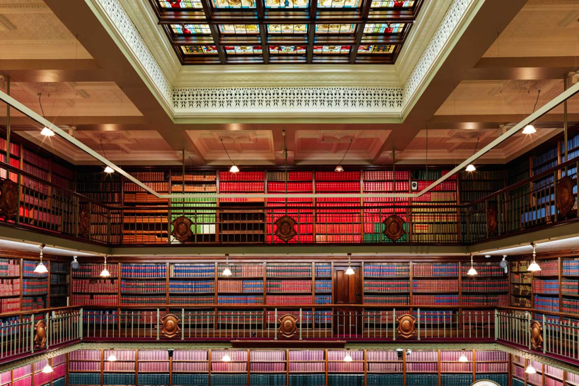 Interior of a grand, multi-level library with balconies, ornate railings and a colorful stained-glass skylight above.