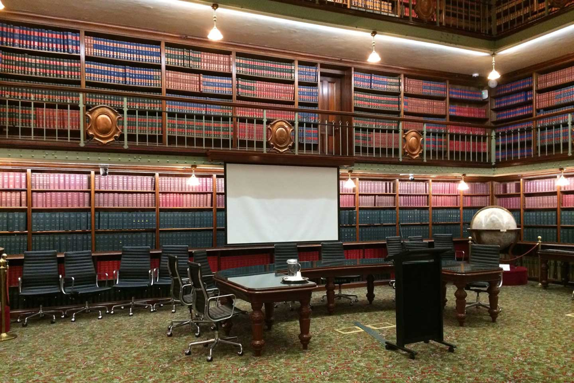 Historic library conference room with tall wooden bookshelves, a projection screen, a globe, and rows of black rolling chairs around dark wooden tables.