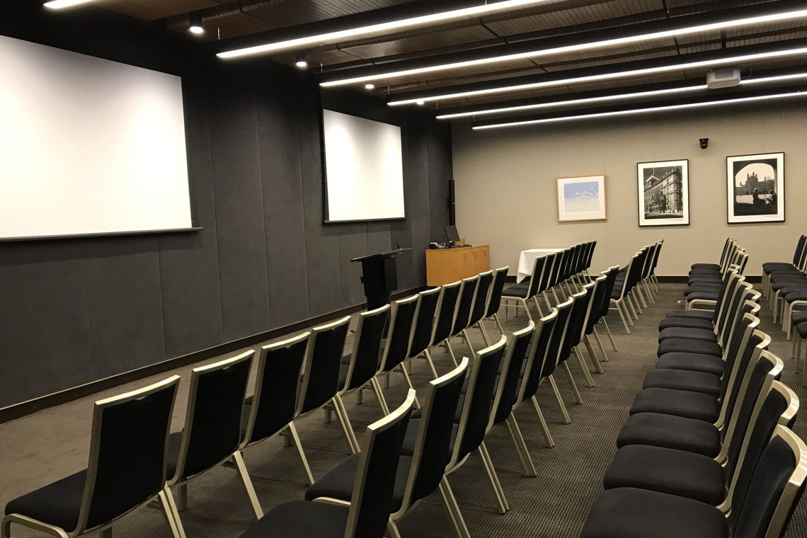 Conference room set with multiple rows of black chairs facing a front stage and white projection screens on dark walls, framed photos on the side wall.