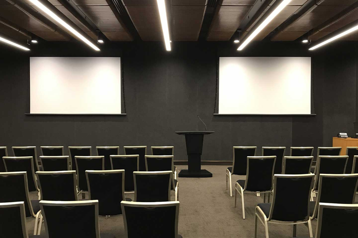 Empty conference or lecture room set up for a presentation: two large white projection screens on a dark wall, a black podium at center, and rows of black chairs facing them.