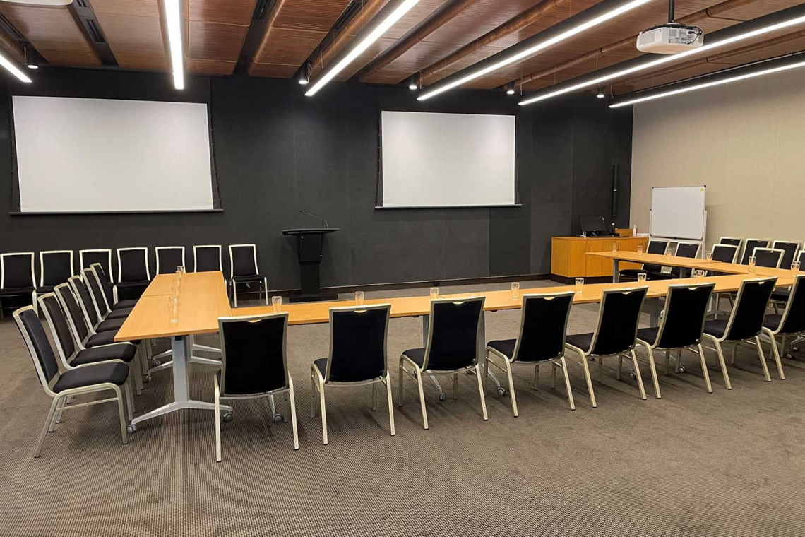 A conference room set up in a U-shaped desk arrangement with black chairs and water glasses, two large projection screens, and a podium at the front.