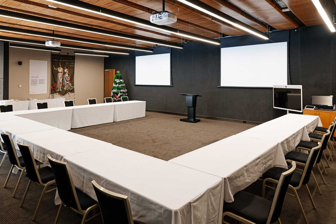 U-shaped conference room setup with white-tablecloth tables, a black podium, two blank projector screens, and a Christmas tree in the corner.