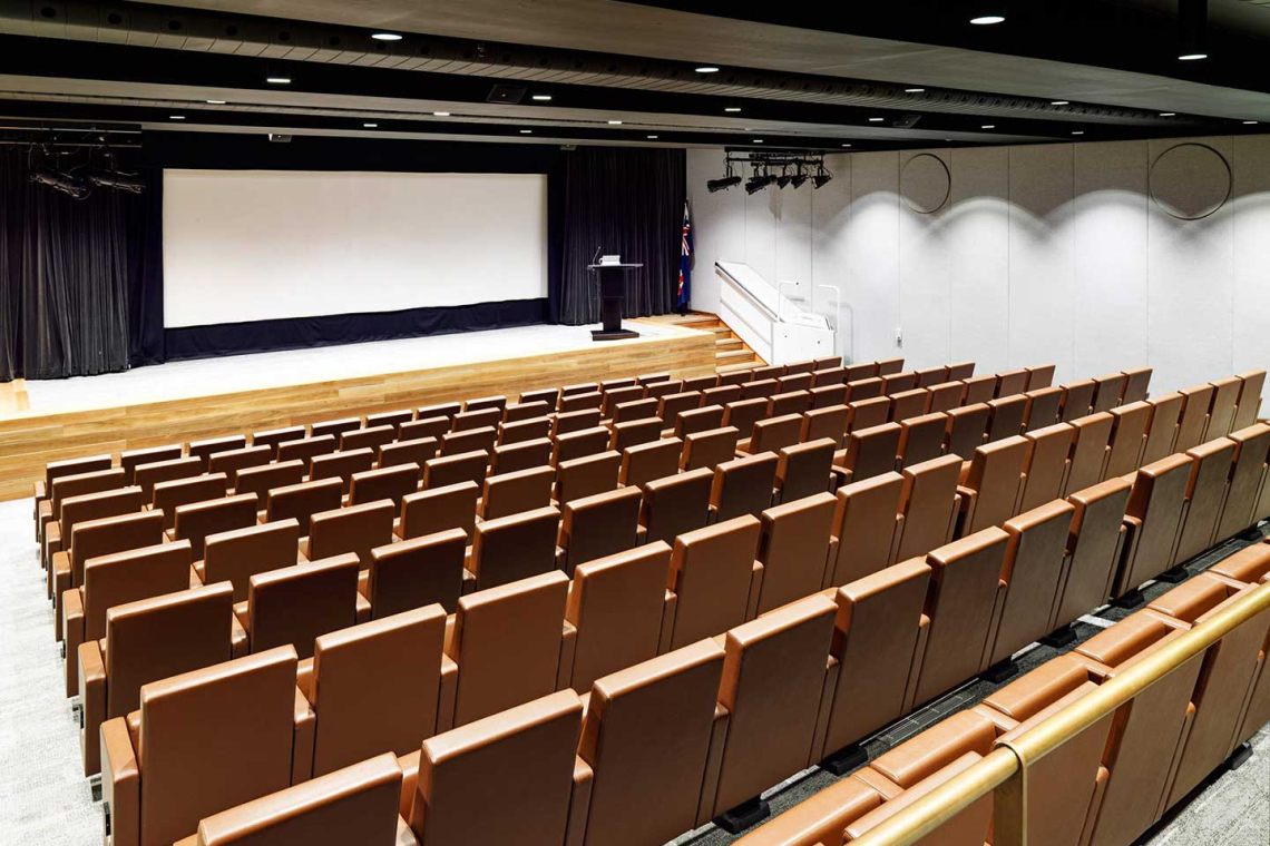 empty auditorium with a wooden stage, a large projection screen, a black curtain backdrop, a podium, and rows of brown upholstered seats; an Australian flag beside the stage