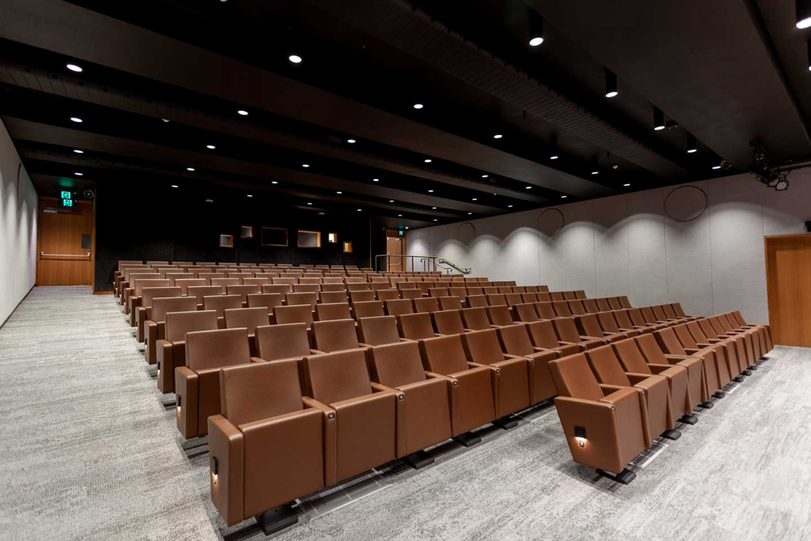 Empty auditorium with rows of brown upholstered seats facing a stage area, lit by ceiling lights.