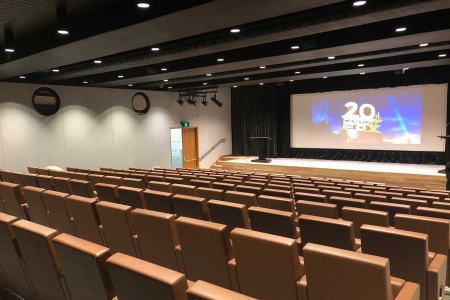 Empty theater with brown upholstered seats facing a stage and a large screen showing the 20th Century Fox logo.