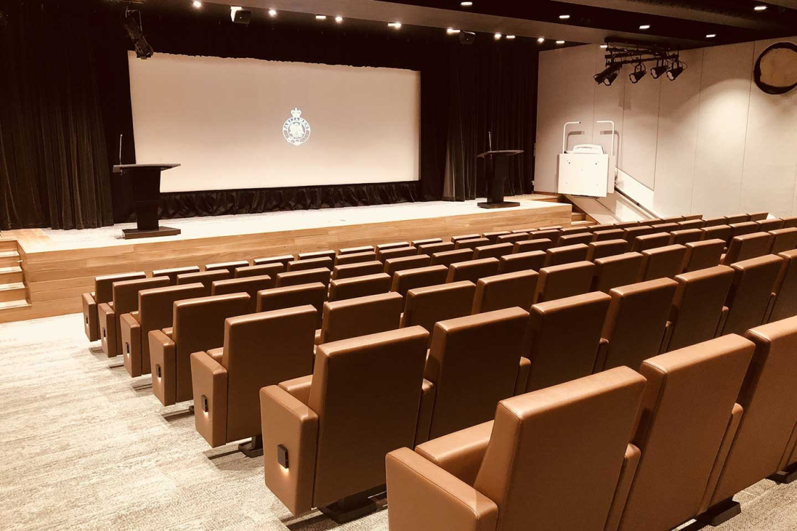 Empty lecture hall with rows of brown seats facing a stage and a large screen.