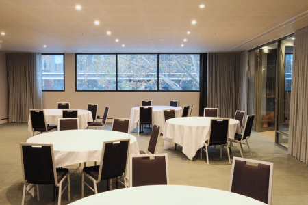Conference room with round tables dressed in white cloths and brown chairs, large windows and soft lighting.