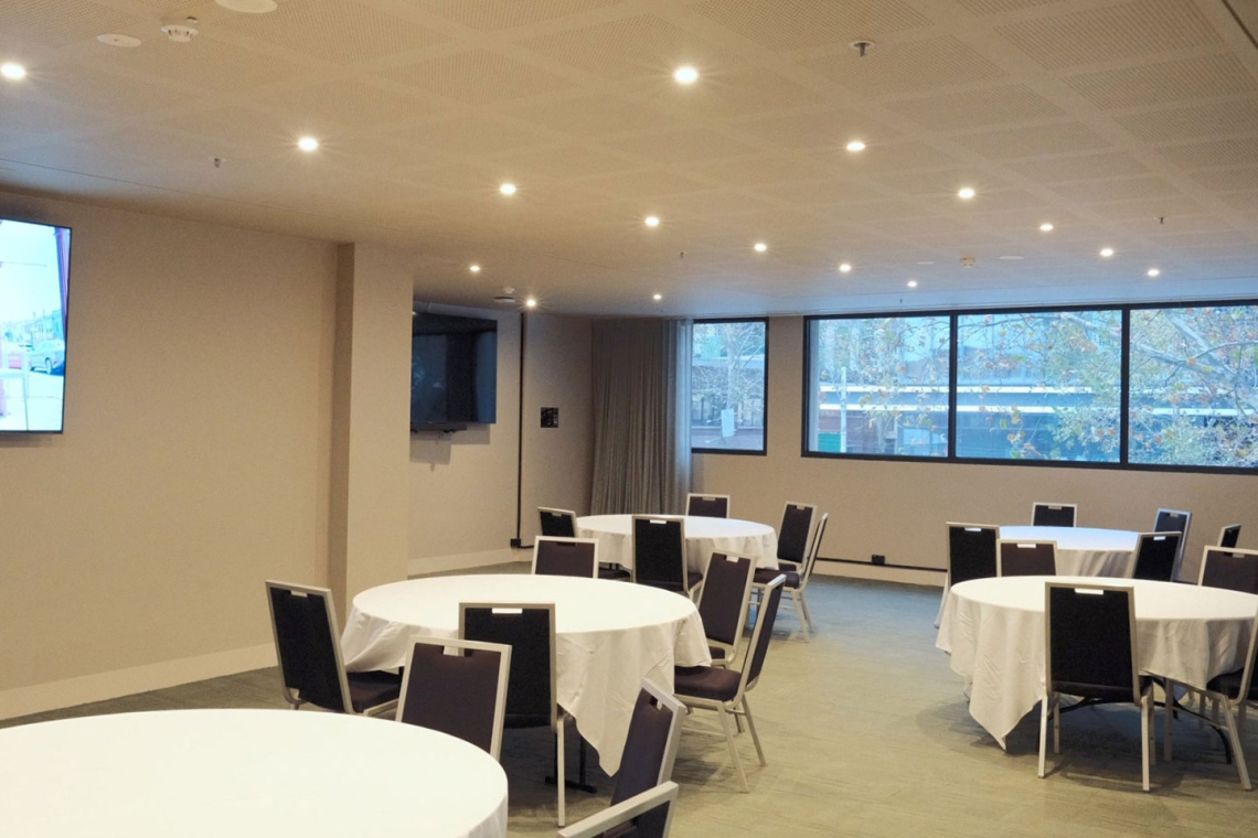 Bright conference room set up for a reception with round tables, white tablecloths, and dark chairs; large windows overlook trees outside.