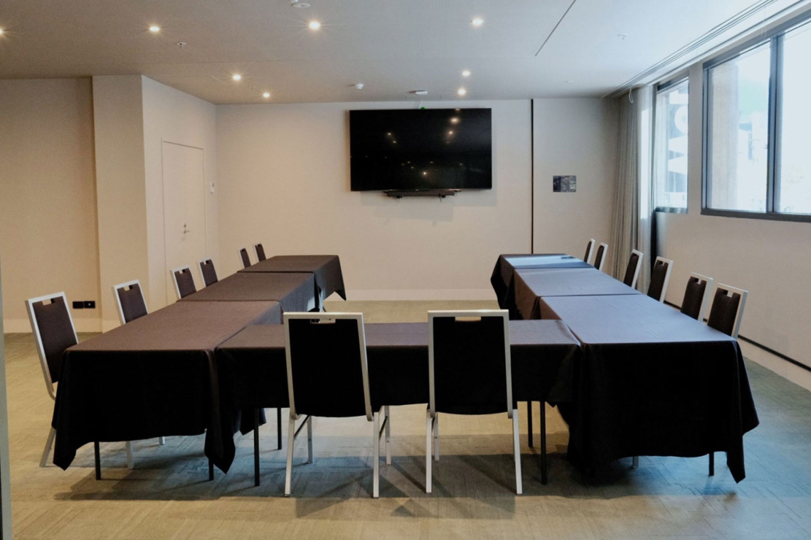 Conference room with black-clothed tables arranged in a U-shape and seating for about a dozen, a wall-mounted flat-screen TV, and large windows on the right.