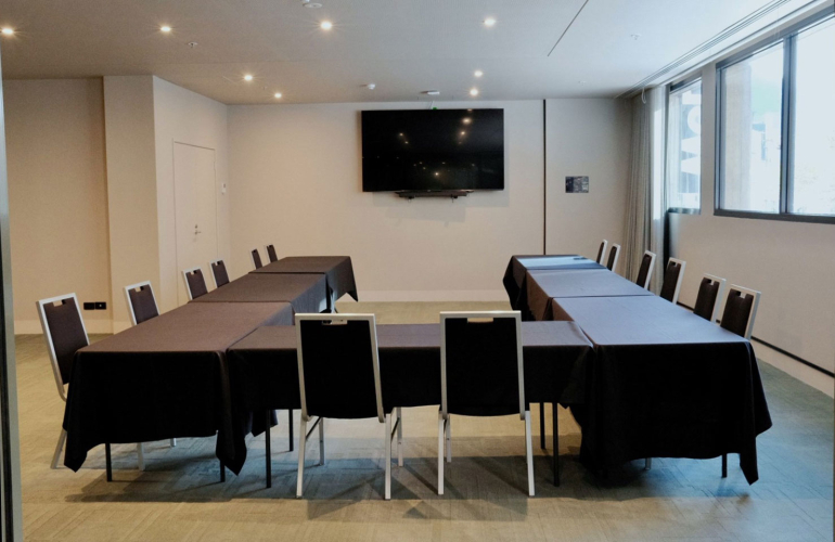 Conference room with black-clothed tables arranged in a U-shape and seating for about a dozen, a wall-mounted flat-screen TV, and large windows on the right.