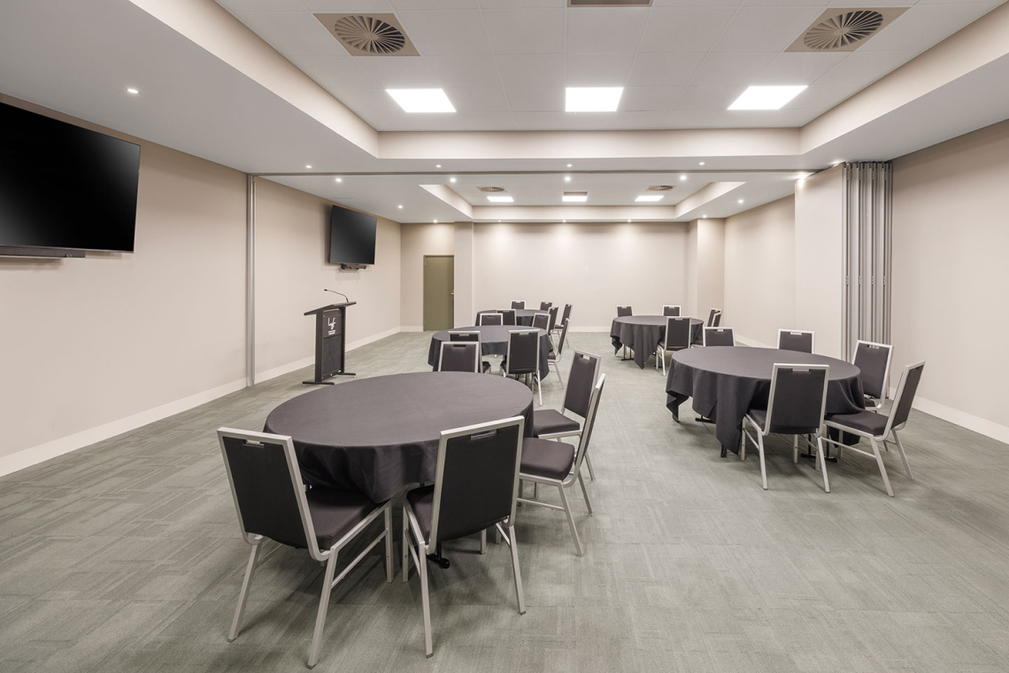 Empty conference room with round tables covered in black cloth and gray chairs, a podium, and wall-mounted TVs in the background.