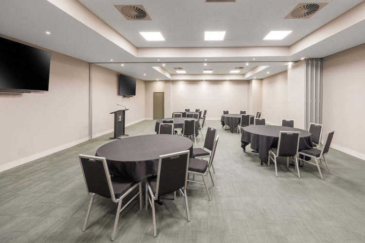 Empty conference room with round tables covered in black cloth and gray chairs, a podium, and wall-mounted TVs in the background.