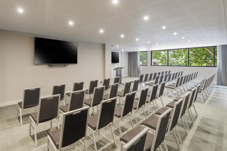 Conference-style room with rows of gray-cushioned chairs facing a lectern and two large wall-mounted TVs, and large windows overlooking greenery.