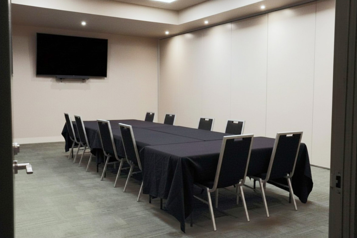 Conference room with a long U-shaped table covered in black cloth, surrounded by chairs, and a large wall-mounted TV at the front.