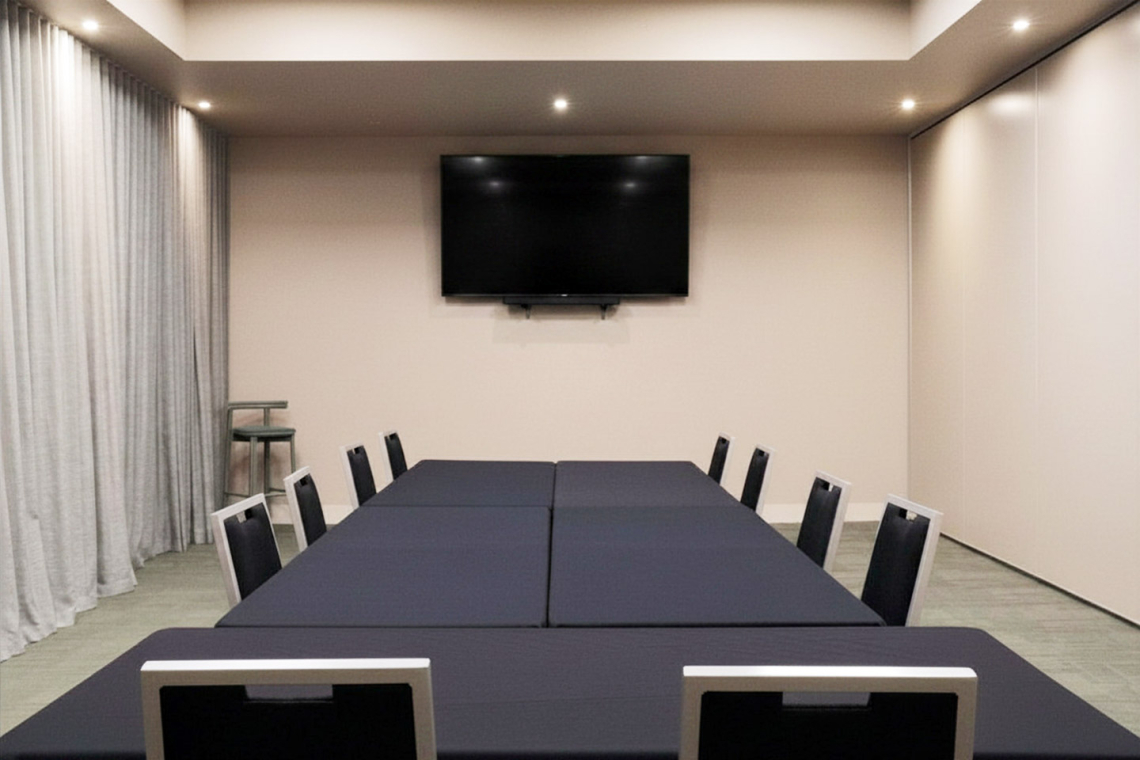 Conference room with a long, dark table and black chairs arranged for a meeting, TV mounted on a beige wall at the far end, soft ceiling lights.