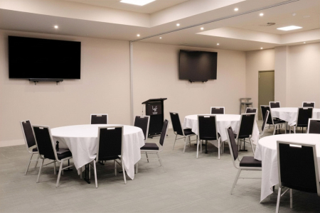 Empty conference room with round tables covered in white cloths, black chairs, a podium at the front, and two wall-mounted TVs.