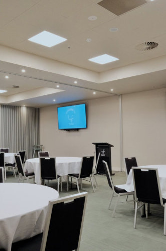 Conference room set up for a seminar with round tables draped in white cloths and black chairs around them, a lectern at the front, and a blue welcome screen on the wall.