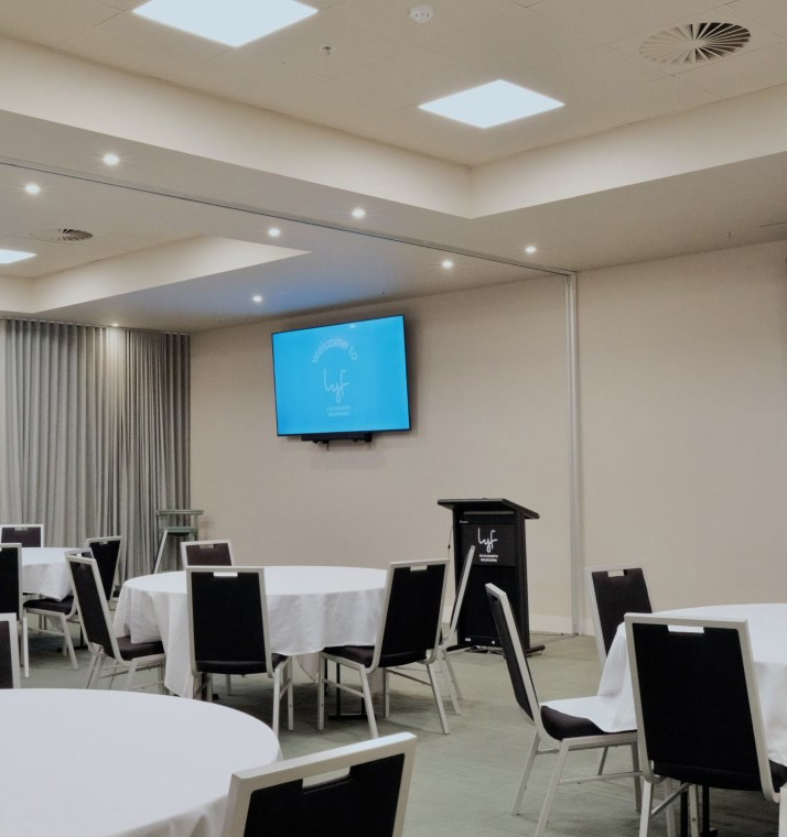 Conference room set up for a seminar with round tables draped in white cloths and black chairs around them, a lectern at the front, and a blue welcome screen on the wall.